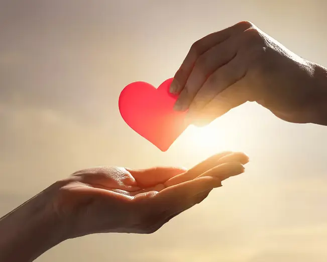 Two hands in front of sunlight, one holding a heart and the other reaching for it, symbolizing generosity and support.