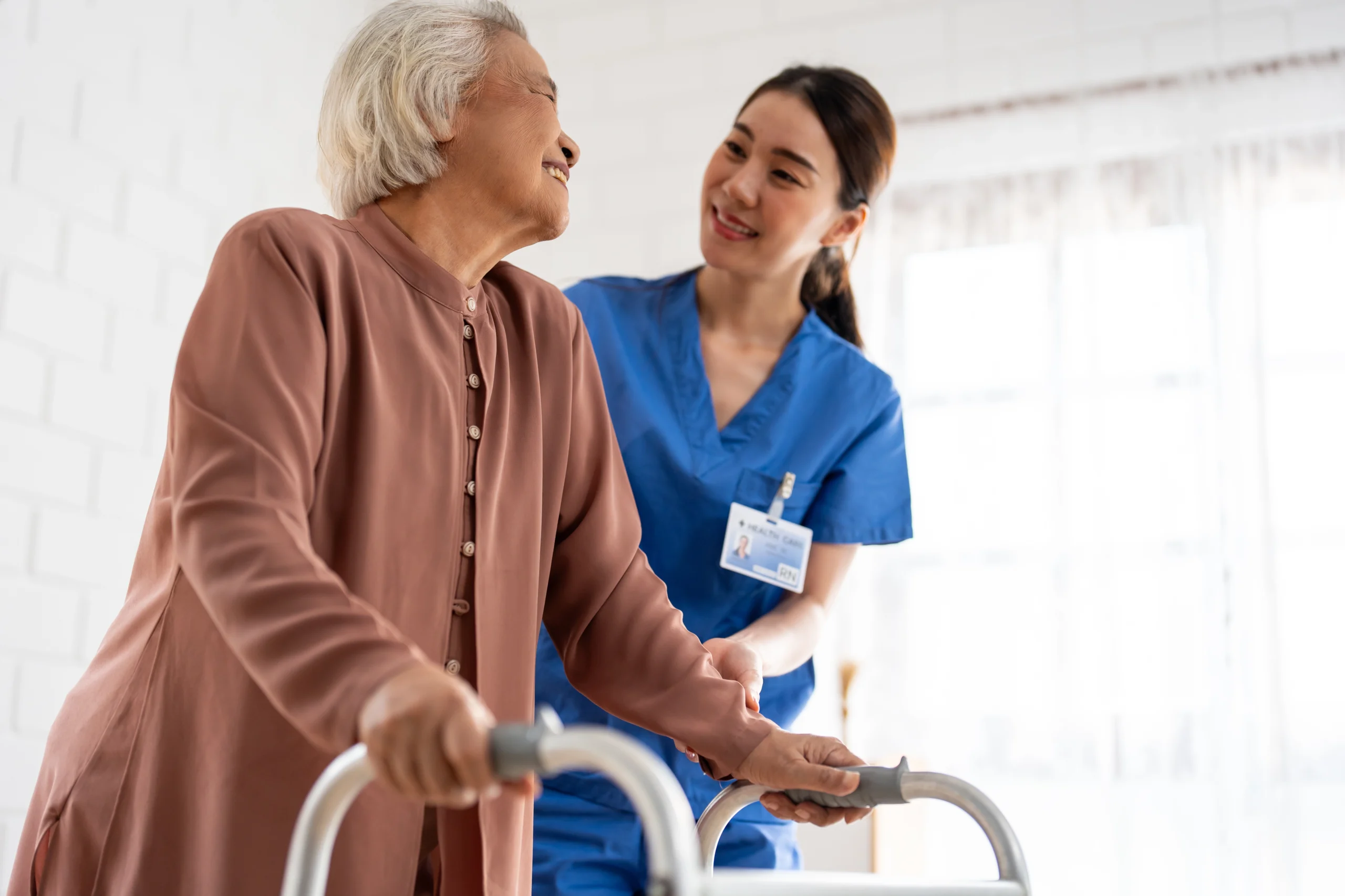 A hospice aide gently holding a patient’s shoulders while assisting them as they walk with a walker.